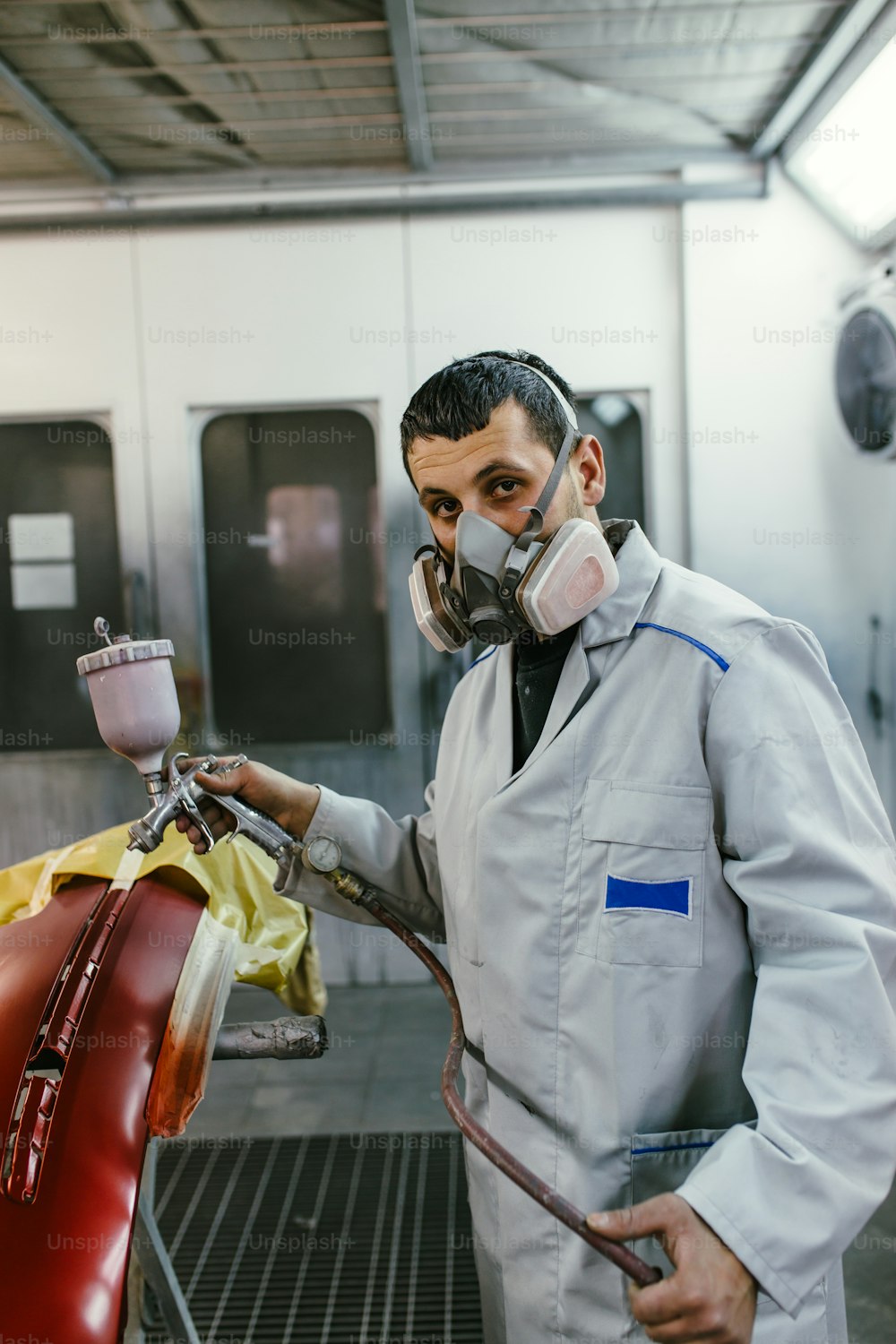 Hombre con ropa protectora y máscara pintando piezas de automóviles con compresor de pulverización. Enfoque selectivo.