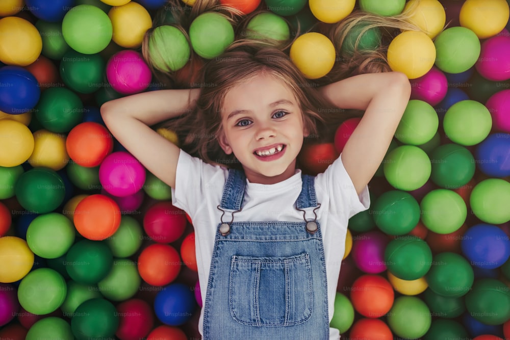 Little pretty girl is having fun outdoor. Playing in children zone in amusement park. Lying in colorful balls and smiling