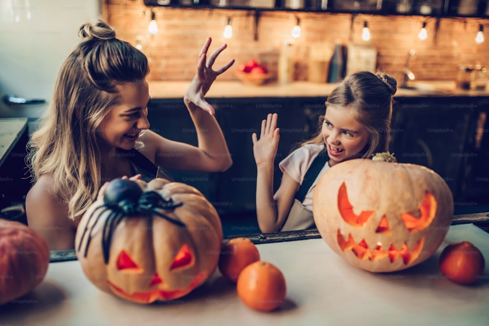 Fröhliches Halloween! Attraktive junge Frau mit ihrer kleinen süßen Tochter bereiten sich auf Halloween in der Küche vor. Mama mit Tochter haben Spaß mit Kürbissen.