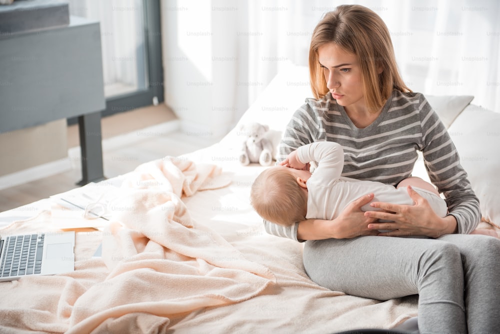 Hush little baby. Troubled mommy is sitting on bed and embracing her child with care. Copy space in left side