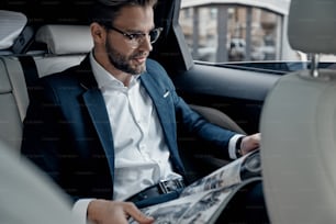 Handsome young man in full suit reading a newspaper while sitting in the car