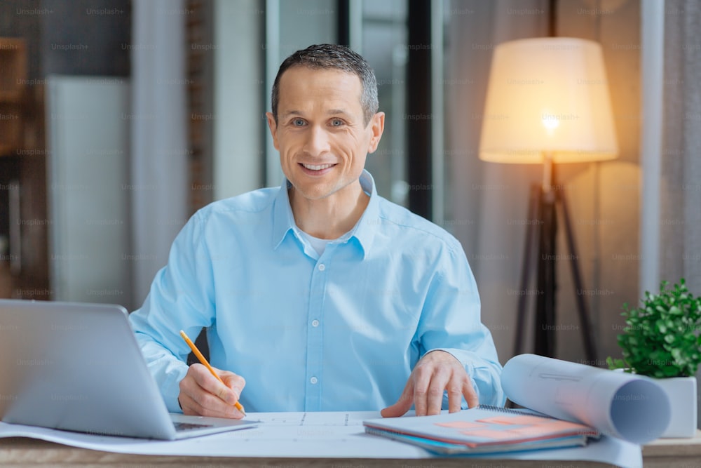 Warm evening. Cheerful male person keeping smile on his face, making sketch while looking straight at camera