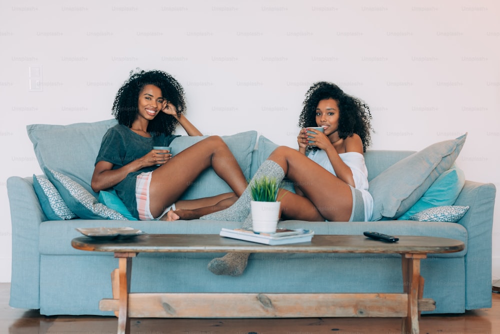 Dos mujeres negras jóvenes felices sentadas en el sofá bebiendo café "n