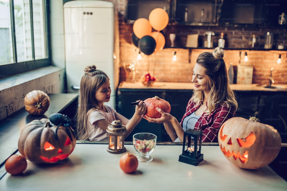 Feliz Dia das Bruxas! Jovem grávida atraente com sua pequena filha fofa estão se preparando para o Halloween na cozinha. Mamãe e filha estão se divertindo com abóboras enquanto pintam.