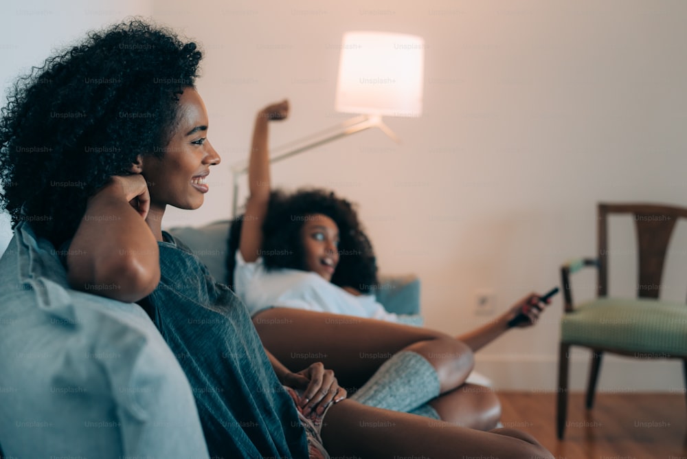 Glückliche junge zwei schwarze Frauen, die auf der Couch liegen und fernsehen