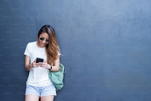 Retrato de estilo de vida al aire libre de una joven asiática bastante sexy en estilo de viaje y gafas sobre fondo de pared gris. Mujer asiática enviando mensajes de texto por teléfono. Espacio de copia disponible. Estilo de vida y tecnología.