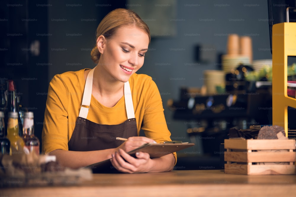 Porträt einer fröhlichen Arbeiterin, die Informationen notiert, während sie die Ellbogen auf dem Tisch in einem gemütlichen Café ausruht. Berufskonzept