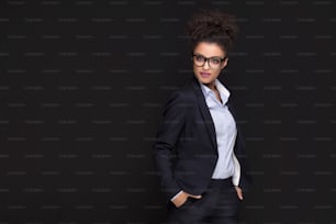 Photo of young african cheerful business woman standing over black background , smiling to the camera.Girl with afro hairstyle wearing eyeglasses.