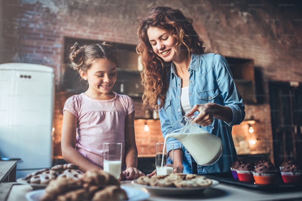 Attractive young woman and her little cute daughter are eating cakes and cookies on kitchen and drinking milk. Having fun together while enjoying "nfreshly baked pastries.