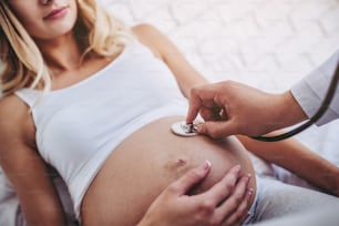 Attraktive schwangere Frau liegt im Bett, während Arzt in der Nähe mit Stethoskop sitzt und Baby hört. Letzte Monate der Schwangerschaft.