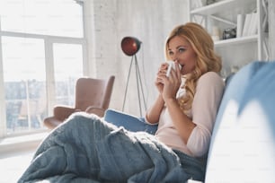 Mulher jovem atraente segurando um copo e olhando para o lado com sorriso enquanto sentado no sofá em casa