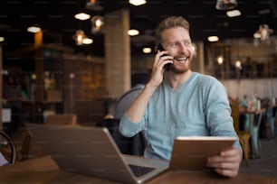 Jovem homem feliz tendo telefonema no café segurando tablet