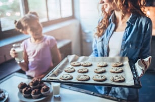 Abgeschnittenes Bild einer attraktiven jungen Frau und ihrer kleinen süßen Tochter essen Kuchen und Kekse in der Küche und trinken Milch. Gemeinsam Spaß haben und dabei "frisch gebackenes Gebäck" genießen.