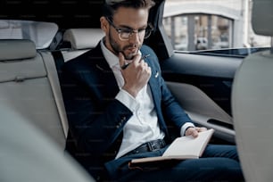 Handsome young man in full suit writing something down in personal organizer while sitting inside of the car