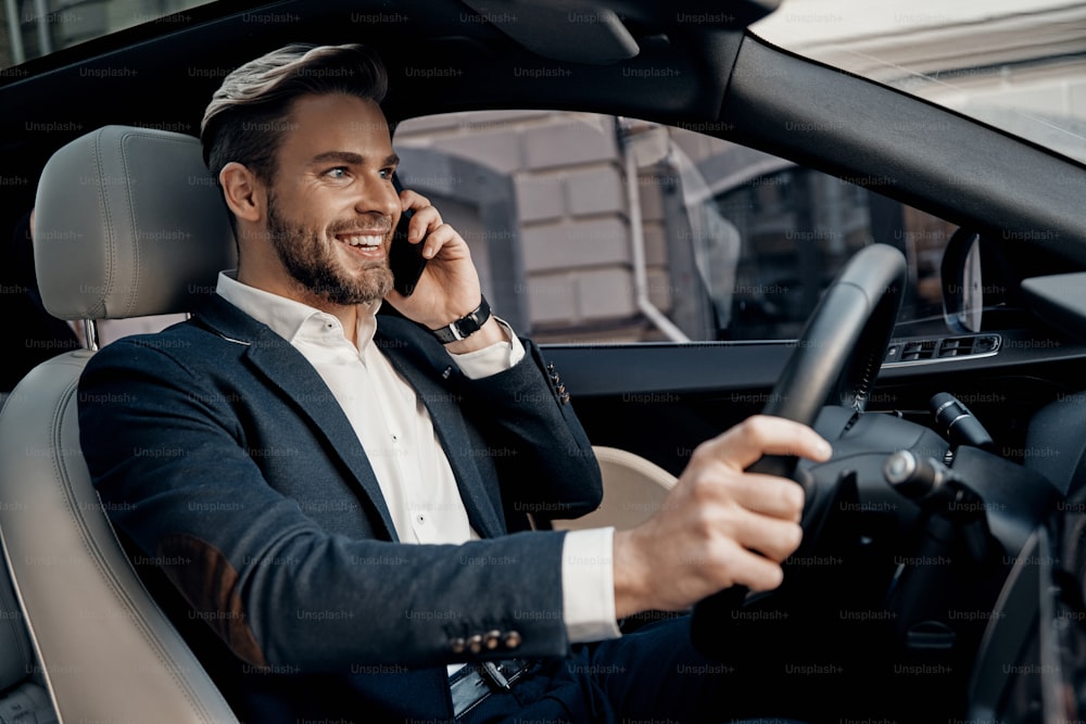 Bel giovane in abbigliamento formale che parla sul suo smartphone e sorride mentre guida un'auto di lusso