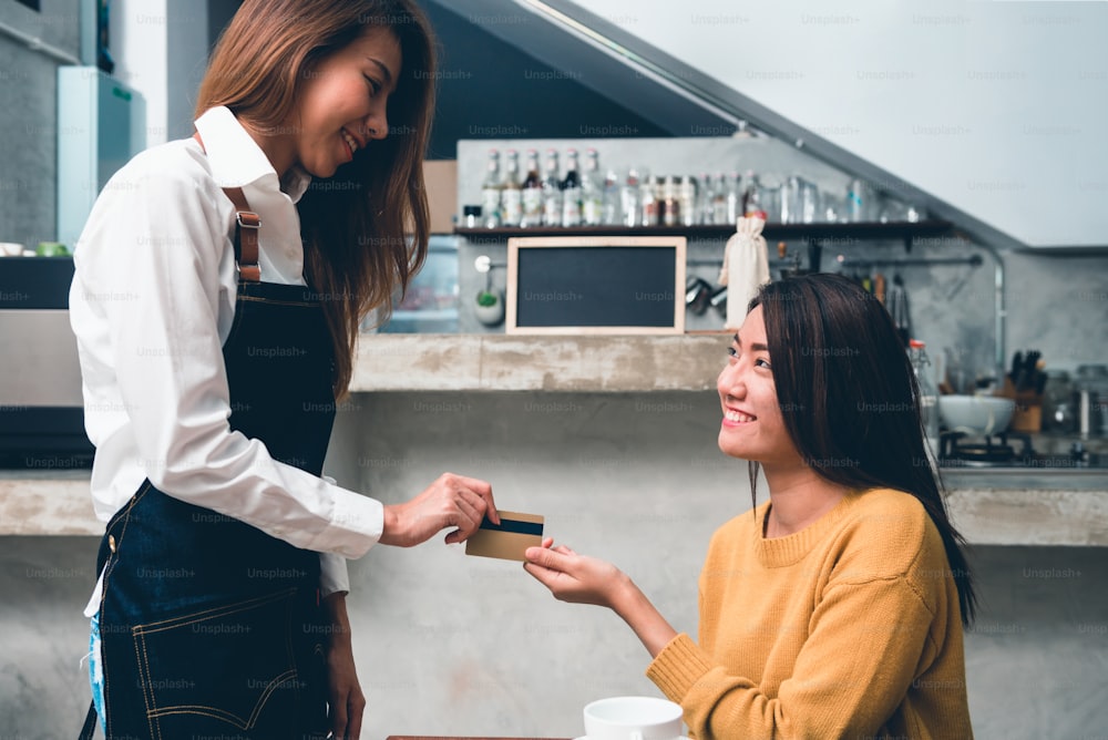 Junge asiatische Frau zahlt per Kreditkarte an den Coffeeshop-Besitzer mit einem Hintergrund der Getränkebartheke in einem kleinen Café. Junge Frau bezahlt ihren Kaffee mit Kreditkarte. Essen und Trinken.