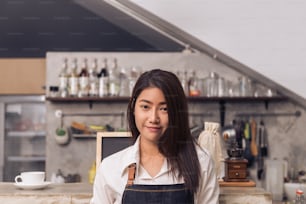 Protrait d’une jeune barista asiatique souriant pour accueillir son client dans un café avec un arrière-plan de comptoir de bar à boissons. Une jeune barista sourit dans son café. Concept de nourriture et de boissons.