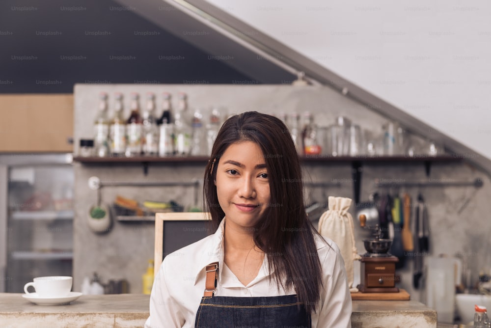 Protrait of young Asian female barista smiling to welcome her customer to coffee shop with a background of beverage bar counter. Young female barista smile in her coffee shop. Food and drink concept.