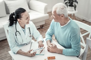 Weekly stock. Mature cute joyful nurse sitting by the table with her patient giving pills to him and smiling.