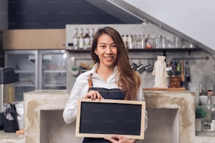 Young Asian female barista in denim apron hold a chalkboard with a beautiful smile in her own coffee shop welcome her customer to try a new coffee menu. Young female barista and her small coffee shop.