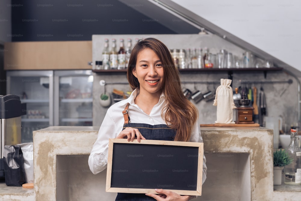 Junge asiatische Barista in Jeansschürze hält eine Tafel mit einem schönen Lächeln in ihrem eigenen Café und begrüßt ihre Kundin, um eine neue Kaffeekarte zu probieren. Junge Barista und ihr kleines Café.