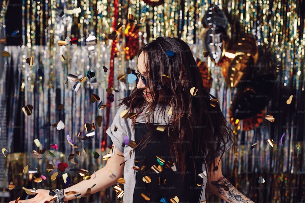 Mulher jovem feliz em roupas da moda celebrando em um fundo de festa cintilante, colorido. Decorações de festa bolas de ouro e prata, balões desfrutando de confetes, preparando-se para abrir confetes.