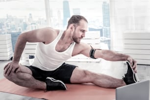 My training. Energetic pleasant active sportsman reaching out leg while putting hand on knee and sitting on mat board