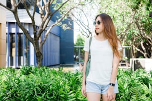 La chica hipster asiática de cabello castaño largo en camiseta blanca en blanco está parada en medio de la calle. Una mujer vestida de calle está de pie sobre un fondo urbano verde. Espacio vacío de maqueta para texto o diseño.
