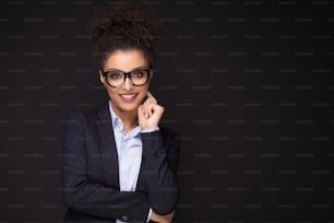 Photo of young african cheerful business woman standing over black background , smiling to the camera.Girl with afro hairstyle wearing eyeglasses.