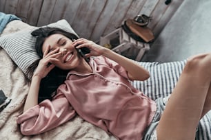 Top view of attractive young woman talking on her smart phone and smiling while lying on the bed at home