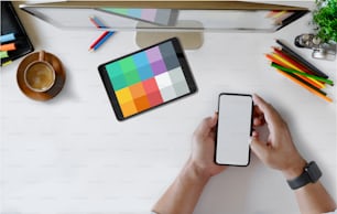 Hombre usando teléfono móvil en la mesa de escritorio blanca de la oficina con computadora de escritorio, suministros de oficina y taza de café. Espacio de trabajo con vista superior y teléfono inteligente con pantalla en blanco