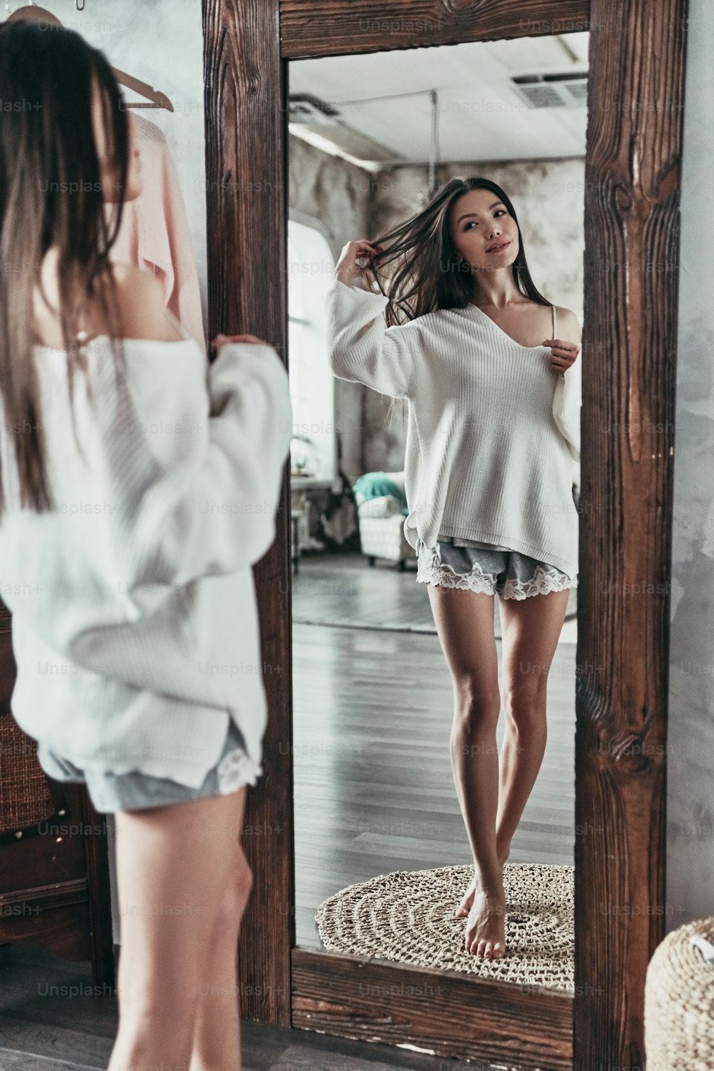 Ganzkörperliches Spiegelbild einer attraktiven jungen Frau, die Hand ins Haar hält und lächelt, während sie zu Hause vor dem Spiegel steht