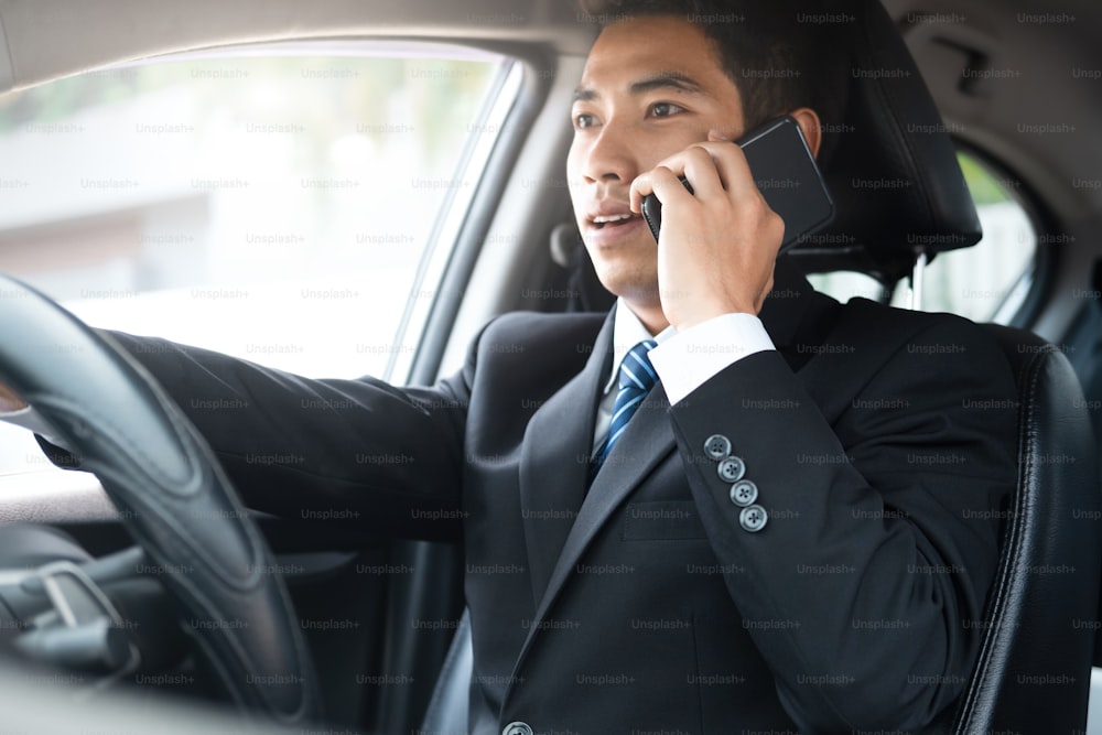 Junger Geschäftsmann spricht auf seinem Smartphone mit ernstem Gesicht, während er das Auto fährt. Wichtiges Telefongespräch. Geschäft im Auto.