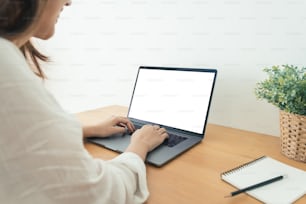 Young Asian woman working using and typing on laptop with mock up blank white screen while at home in office work space. Businesswoman working from home via portable computer. Enjoying time at home.