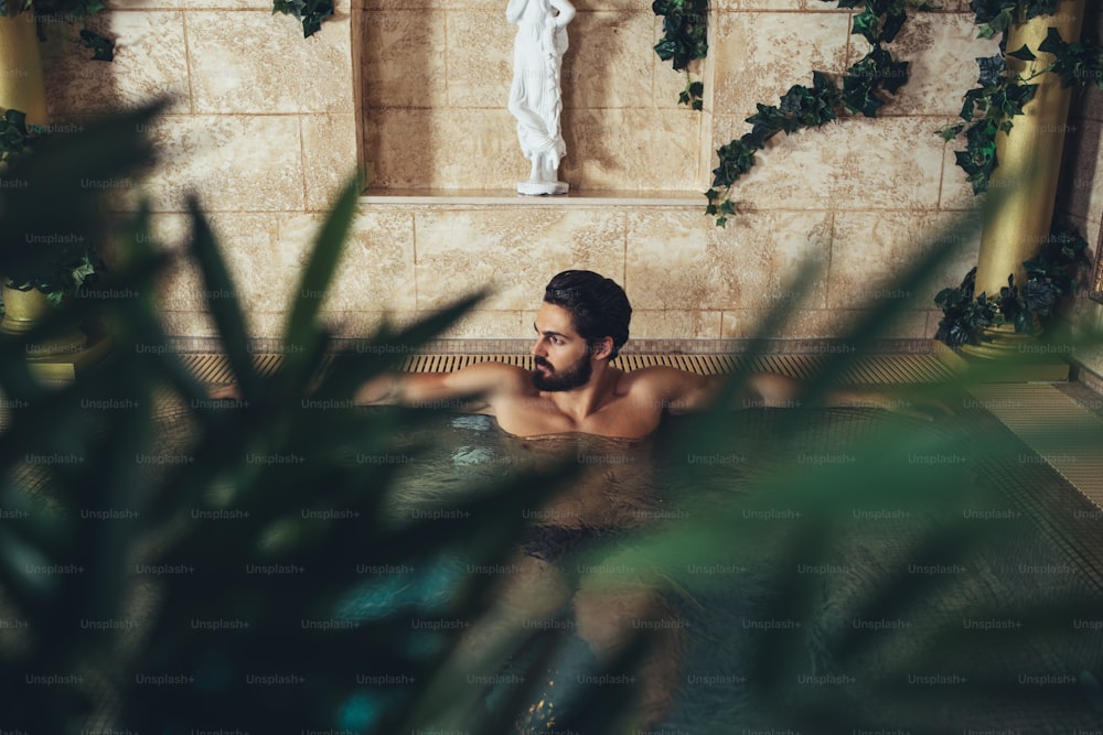 Young handsome bearded man enjoying and relaxing in exclusive spa resort. He sitting in hot tub and drinking wine.