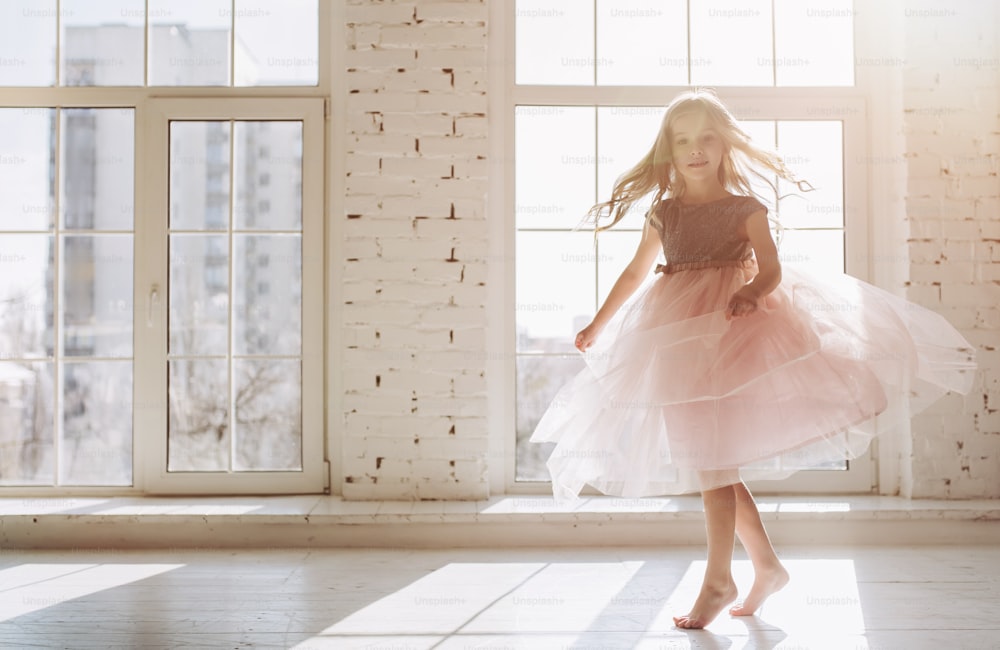 La bambina carina in un bel vestito balla in una stanza soleggiata leggera