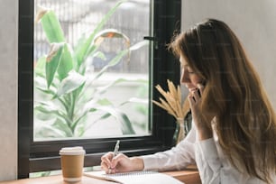 Young business woman in white dress sitting at table in cafe and writing in notebook. Asian woman talking smartphone and cup of coffee. Freelancer working in coffee shop. Student learning online.