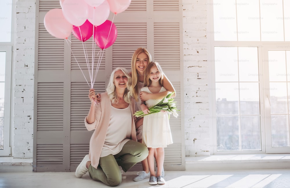 Little cute girl, her attractive young mother and charming grandmother are standing with air balloons and flowers in light room. Women's generation. International Women's Day. Happy Mother's Day.