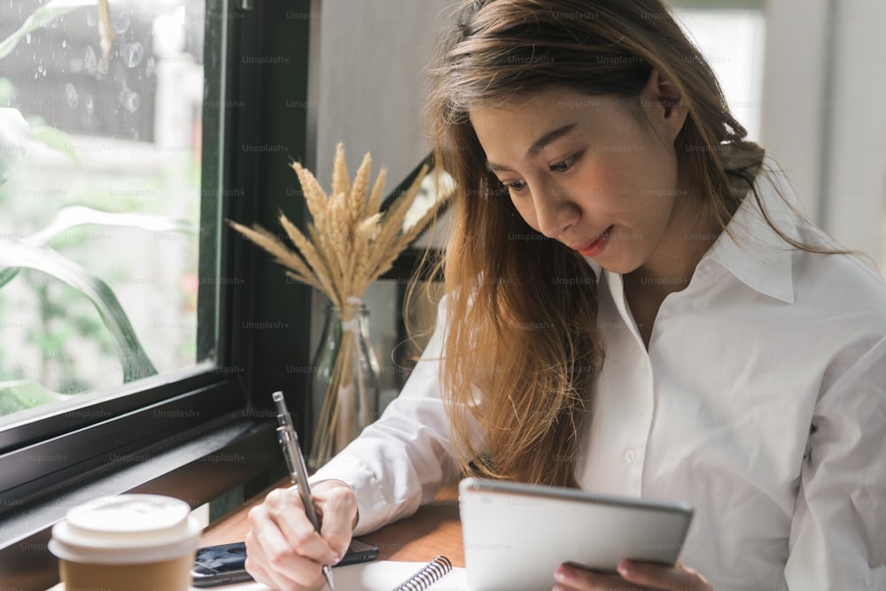 Joven mujer de negocios en vestido blanco sentada en la mesa en la cafetería y escribiendo en el cuaderno. Mujer asiática usando tableta y taza de café. Freelancer trabajando en cafetería. Aprendizaje de estudiantes en línea.