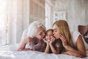 Little cute girl, her attractive young mother and charming grandmother are lying on bed and spending time together at home. Women's generation. International Women's Day. Happy Mother's Day.