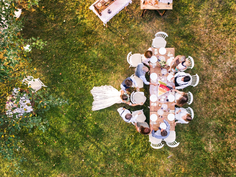 Recepción de bodas al aire libre en el patio trasero. Novia y novio con una familia de pie alrededor de la mesa, saludando. Vista aérea.