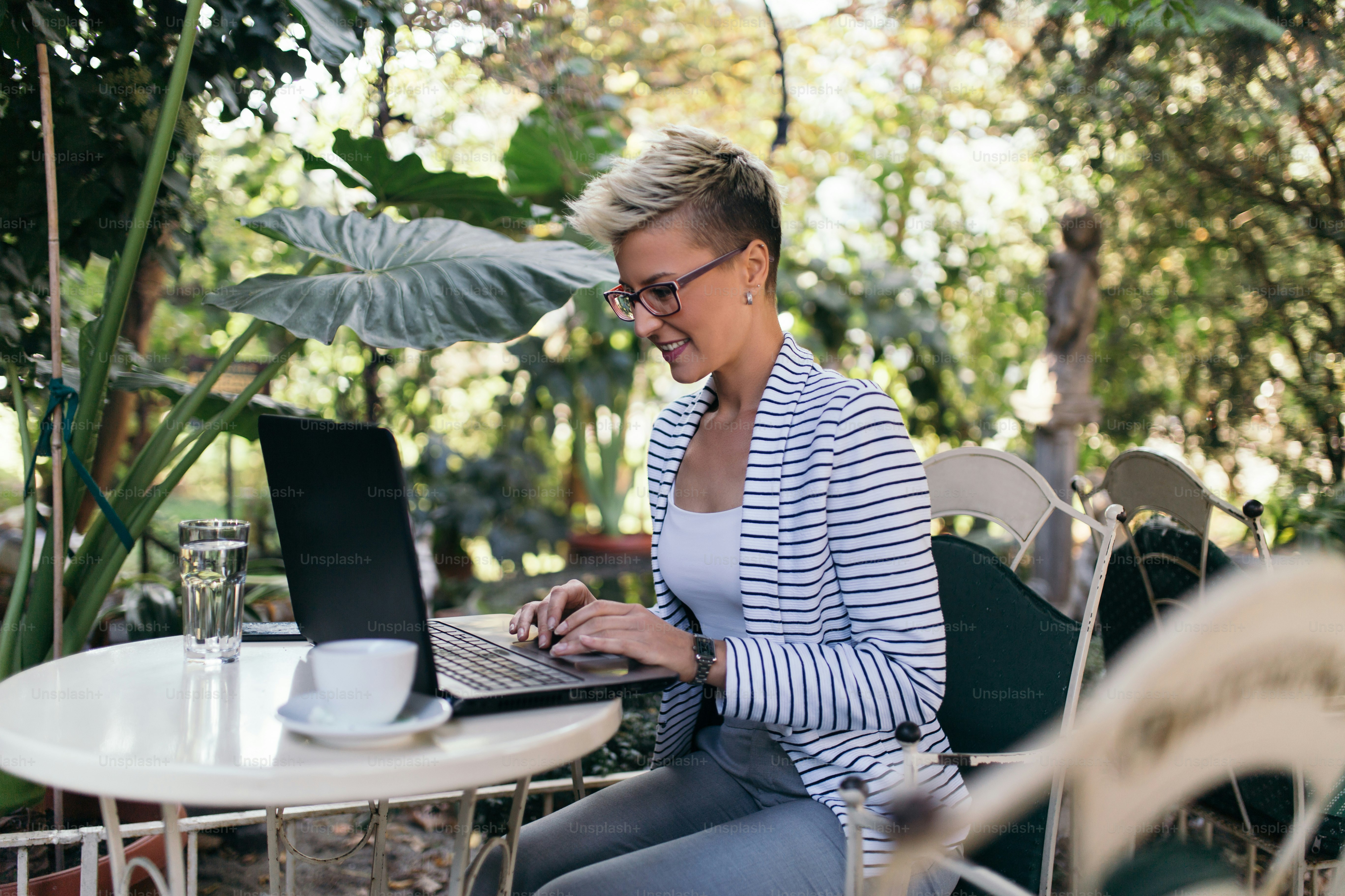 Jeune femme heureuse assise dans un beau jardin comme un café, un bar ou un restaurant et faisant quelque chose sur son ordinateur portable.