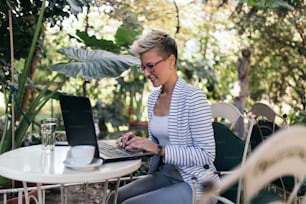 Young happy woman sitting in beautiful garden like cafe bar or restaurant and doing something on her laptop computer.