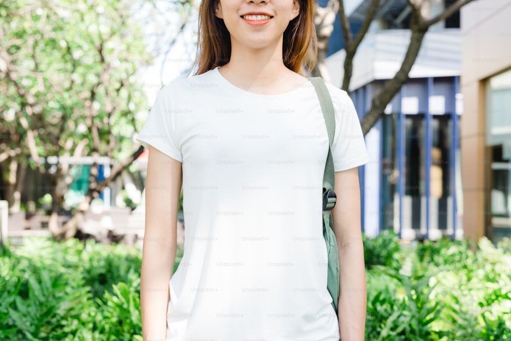 Asiatisches Hipster-Mädchen lange braune Haare in weißem leerem T-Shirt steht mitten auf der Straße. Eine Frau in Streetwear steht auf einem grünen urbanen Hintergrund. Leerer Mock-up-Bereich für Text oder Design.