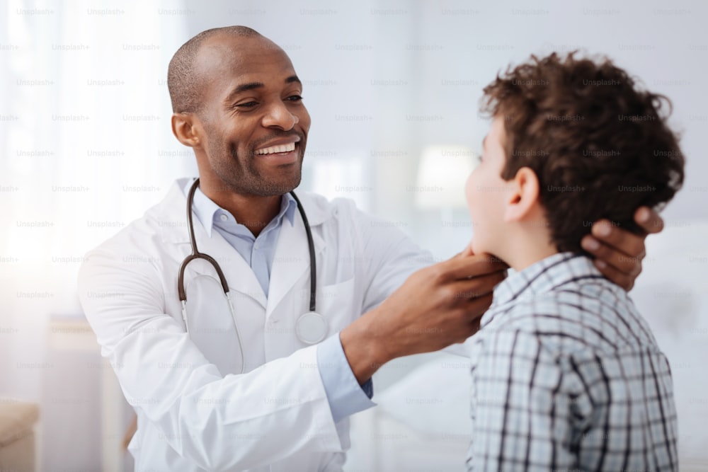 Schmerzen im Hals. Hübscher amerikanischer Afro-Arzt, der den Jungen ansieht, während er lächelt und seine Kehle überprüft