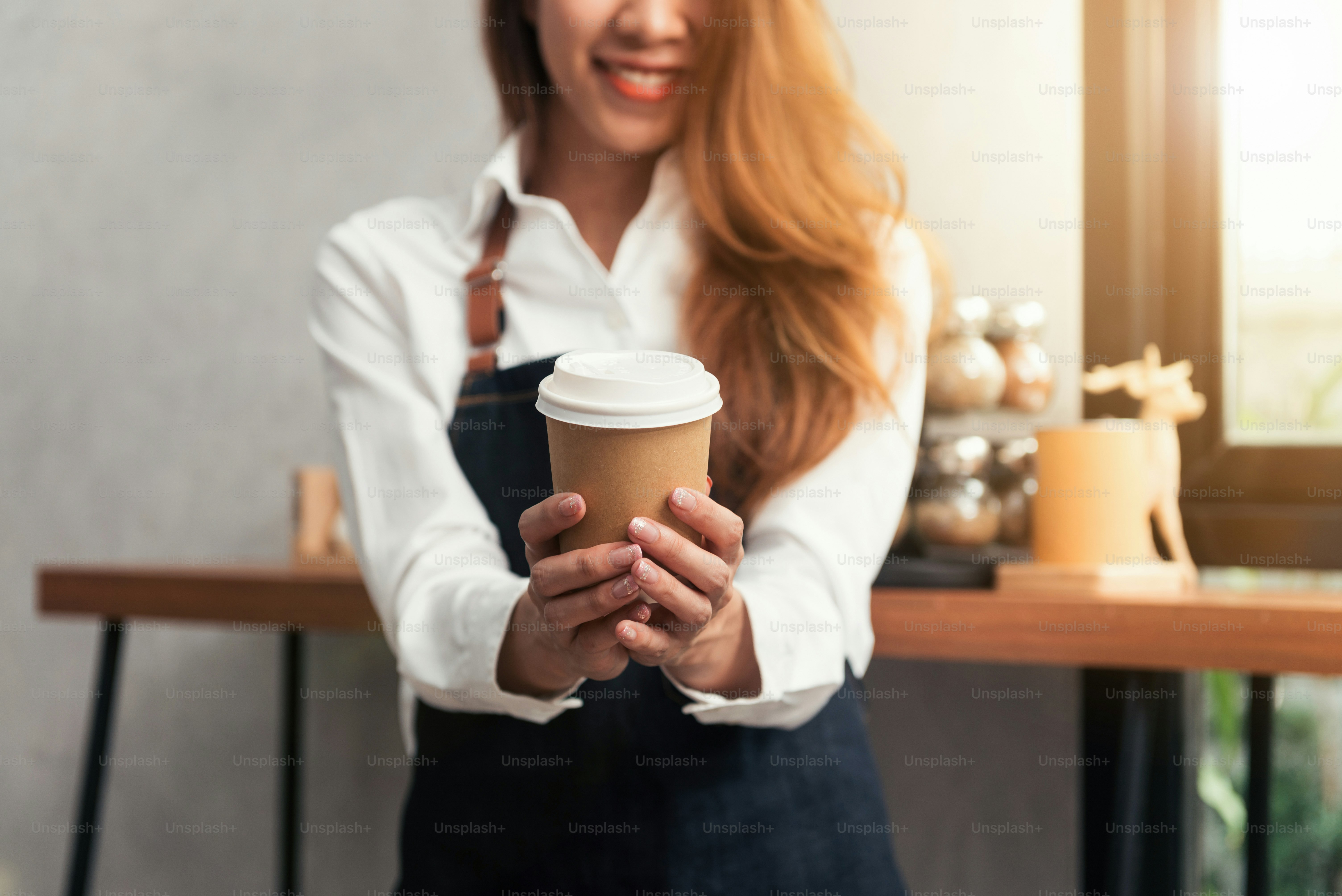 Close up of a young Asian female barista hold a cup of coffee serving to her customer with smile surrounded with bar counter background. Young female barista and her small shop. Food and drink concept