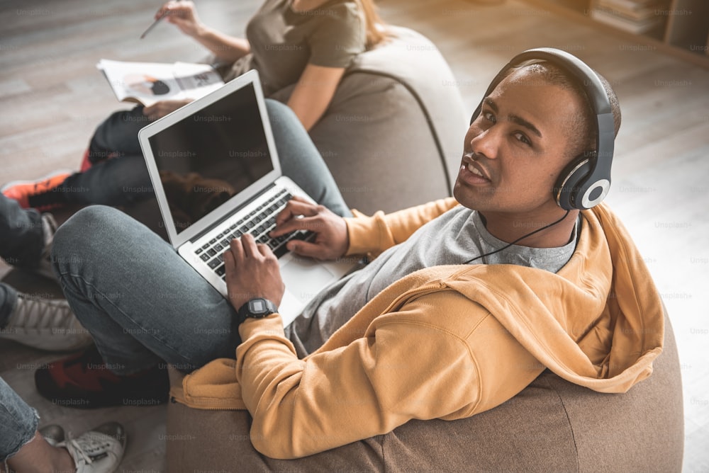 Portrait of african guy with headphones on head sitting on flexible chair with laptop on his knees. He is staring at camera with serenity. Coworkers on background