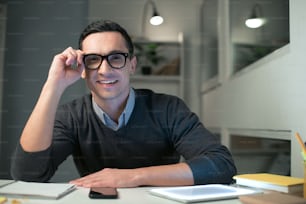Ready to work. Gay vigorous male programmer touching glasses while sitting at the table and looking at the camera