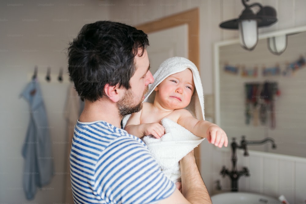 Vater mit einem unglücklichen Kleinkind, eingewickelt in ein Handtuch in einem Badezimmer zu Hause. Vaterschaftsurlaub.