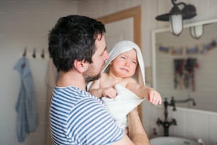 Father with an unhappy toddler child wrapped in a towel in a bathroom at home. Paternity leave.